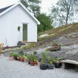 En vit stuga med gröna detaljer och ett bord med stolar på en grusad uteplats. Blommor och växter i krukor pryder den steniga sluttningen.