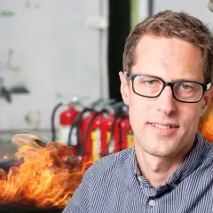 Man med glasögon framför brandsläckare och flammor. En påminnelse om vikten av boverkets byggregler och brandskydd.