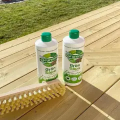 Två flaskor Grumme Grön Såpa och en borste ligger på en träaltan, redo att behandla altanen med såpa för en ren och fräsch yta.
