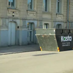 Stor container för avfall står på gatan framför en byggnad. Den är märkt med "Kasta" och kontaktuppgifter.