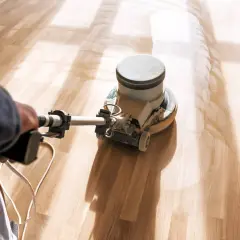 En person slipar ett ljust trägolv med en maskin. Dammet virvlar upp och skapar ett mönster på golvet.