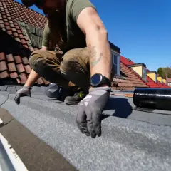 Man spikar takpapp på ett tak med tegelpannor. Solen skiner och himlen är blå.