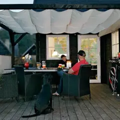 En mysig uteplats med segelduk som tak. Ett par sitter vid ett bord med en hund, redo för en trevlig kväll.