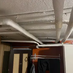 Isolerade rör löper längs det vitmålade svall taket i ett utrymme med träpanel och en dörr.