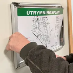 En hand justerar en utrymningsplan på väggen. Planen visar byggnadens planlösning och instruktioner för utrymning, vilket är viktigt enligt utrymningslarm regler.