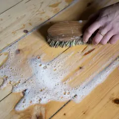 En hand skurar ett trägolv med en borste och såpvatten. Ett enkelt såpvatten recept kan ge ett skinande rent resultat.