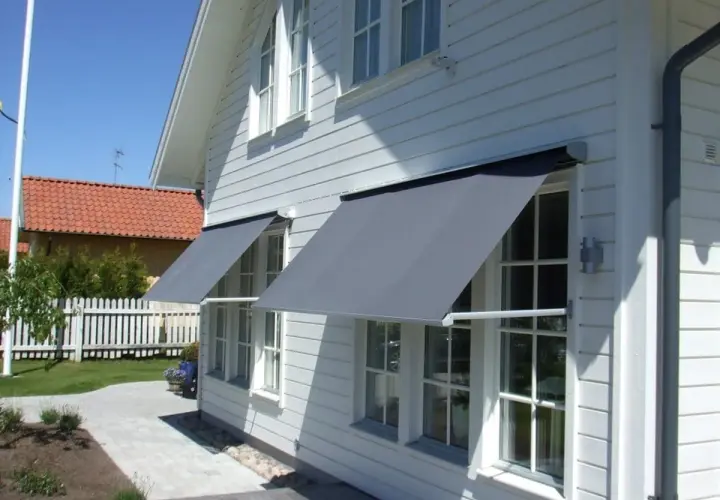 Grå markiser monterade på ett vitt trähus ger skugga. En vacker trädgård med gångväg syns i bakgrunden.