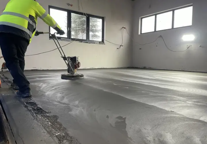 Arbetare polerar nylagd betong med en maskin. Golvet är fortfarande fuktigt och reflekterar ljuset. En del av ballast betong syns längs kanten.