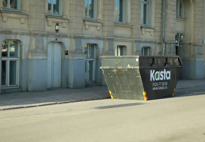 Stor container för avfall står på gatan framför en byggnad. Den är märkt med "Kasta" och kontaktuppgifter.
