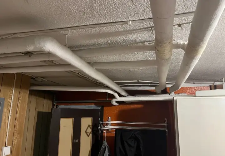 Isolerade rör löper längs det vitmålade svall taket i ett utrymme med träpanel och en dörr.