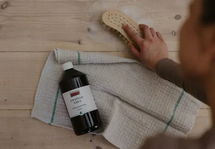 En hand skrubbar trägolv med en borste och linoljesåpa. Flaskan med linoljesåpa är märkbar, men var försiktig, den är brandfarlig.