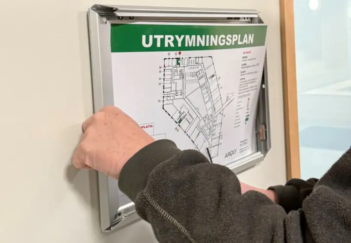En hand justerar en utrymningsplan på väggen. Planen visar byggnadens planlösning och instruktioner för utrymning, vilket är viktigt enligt utrymningslarm regler.