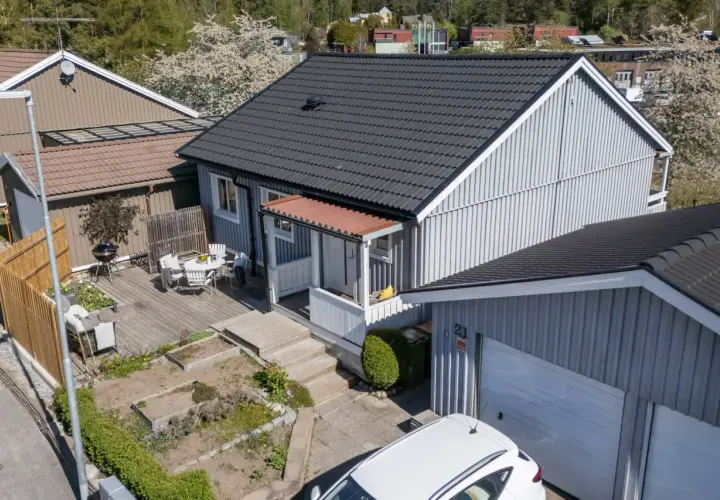 Ett charmigt hus med nummer bbr 21. Trädgårdsmöbler på altanen, en grill och en vit bil framför garaget.