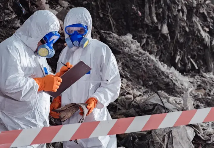 Två personer i skyddsdräkter och gasmasker inspekterar avfall. De följer asbestsanering regler för att säkerställa en trygg miljö.