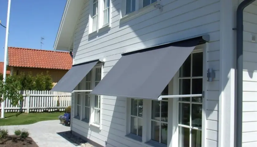 Grå markiser monterade på ett vitt trähus ger skugga. En vacker trädgård med gångväg syns i bakgrunden.
