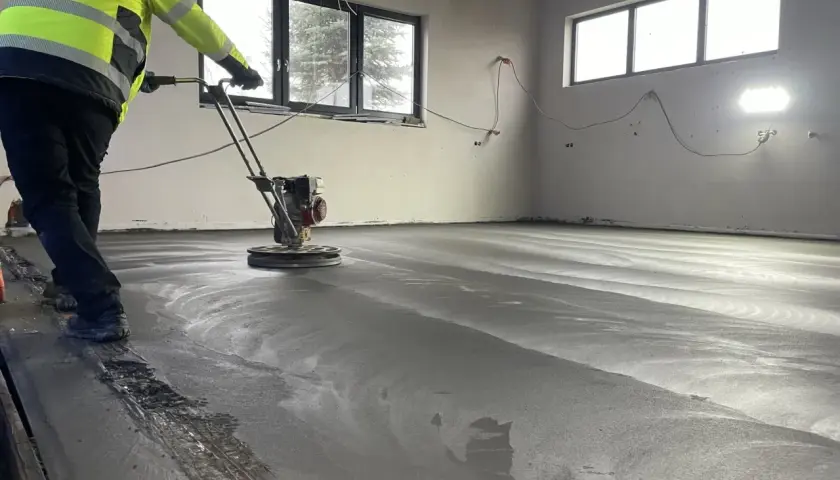 Arbetare polerar nylagd betong med en maskin. Golvet är fortfarande fuktigt och reflekterar ljuset. En del av ballast betong syns längs kanten.