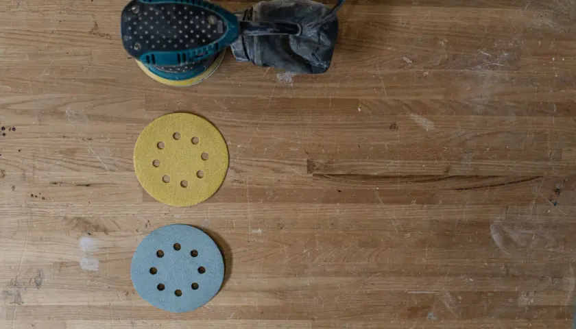 Verktyg för att slipa parkettgolv: en slipmaskin och två slippapper, ett gult och ett blått, ligger på ett slitet trägolv.