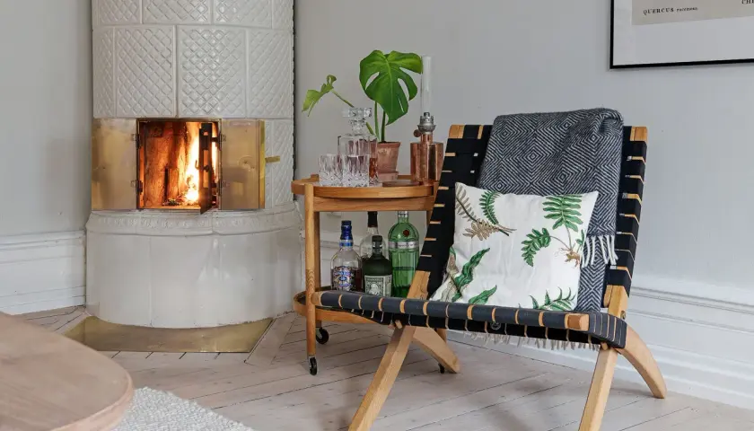 Vacker kakelugn med tänd eld. En stol med pläd och kudde står bredvid ett serveringsbord med drycker. Tänk på renovera kakelugn pris för en mysig atmosfär.