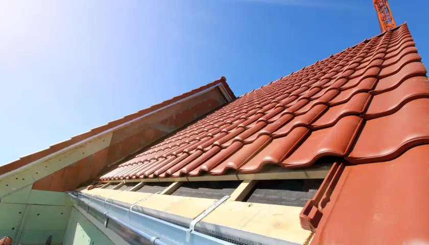 Taktegel i rött läggs på ett hus under uppbyggnad. En kran syns i bakgrunden, redo att lyfta material för framtida byggnadsintegrerade solceller.
