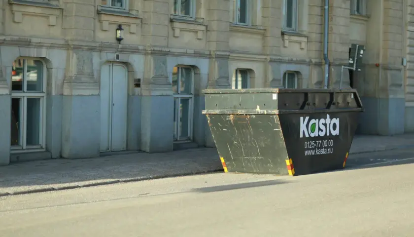 Stor container för avfall står på gatan framför en byggnad. Den är märkt med "Kasta" och kontaktuppgifter.