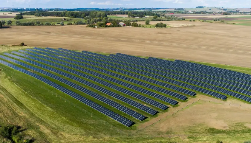 En stor solcellspark på åkermark, med rader av solpaneler som sträcker sig över grönskande och brun jordbruksmark.