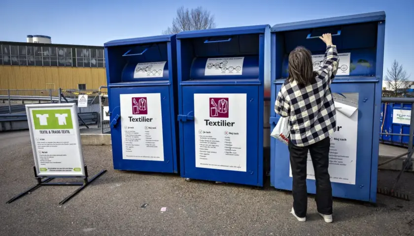 Person lämnar kläder i blå container för textilier återvinning. Skylt visar vad som får lämnas.