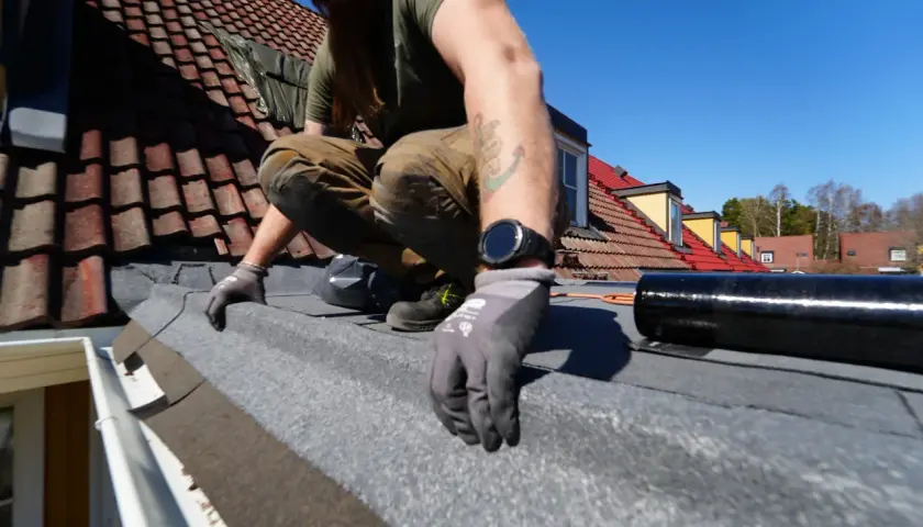 Man spikar takpapp på ett tak med tegelpannor. Solen skiner och himlen är blå.