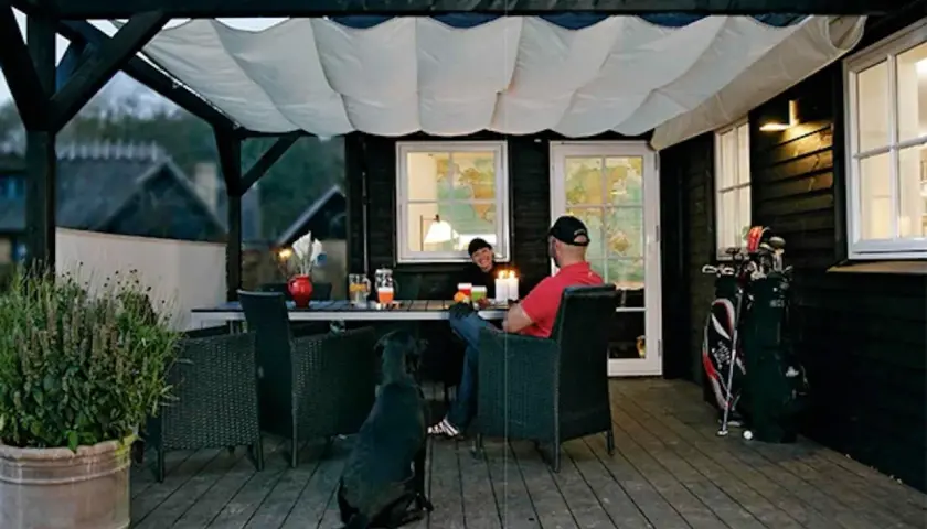 En mysig uteplats med segelduk som tak. Ett par sitter vid ett bord med en hund, redo för en trevlig kväll.