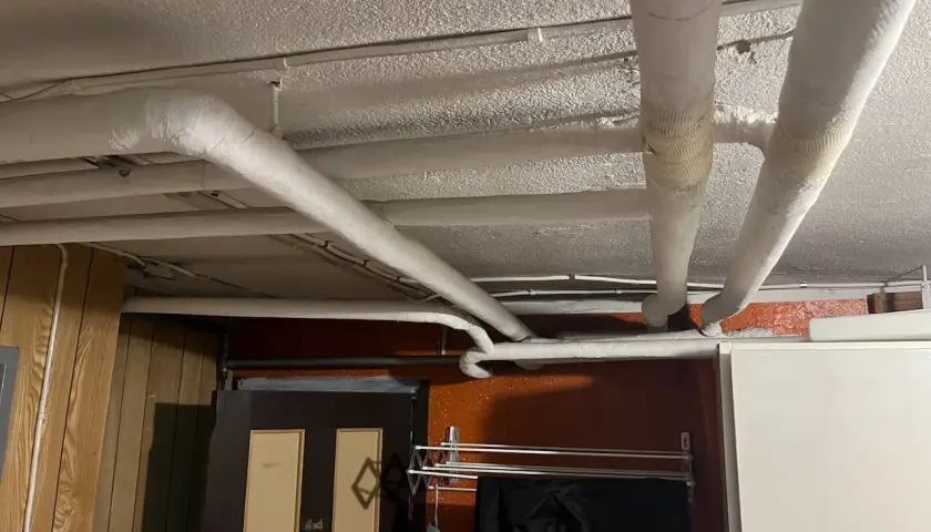 Isolerade rör löper längs det vitmålade svall taket i ett utrymme med träpanel och en dörr.