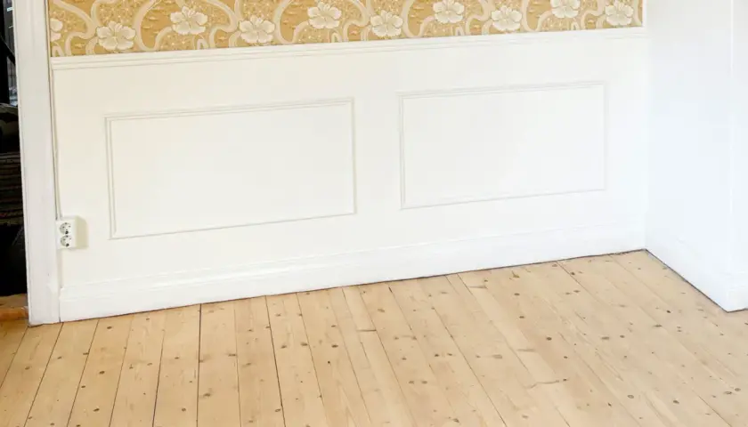 Ett rum med en blommig tapet, vitmålad panel och ett trägolv som väntar på att behandlas med såpa med linolja.