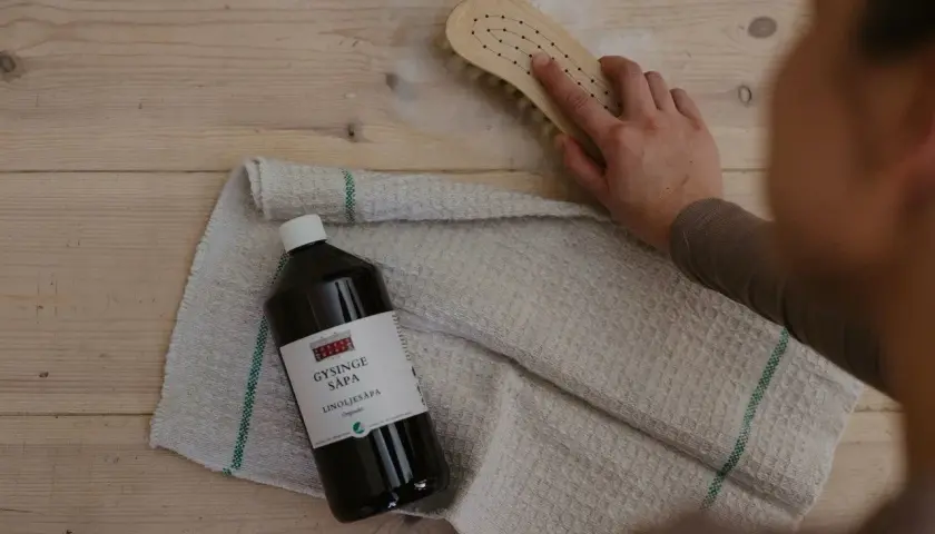 En hand skrubbar trägolv med en borste och linoljesåpa. Flaskan med linoljesåpa är märkbar, men var försiktig, den är brandfarlig.