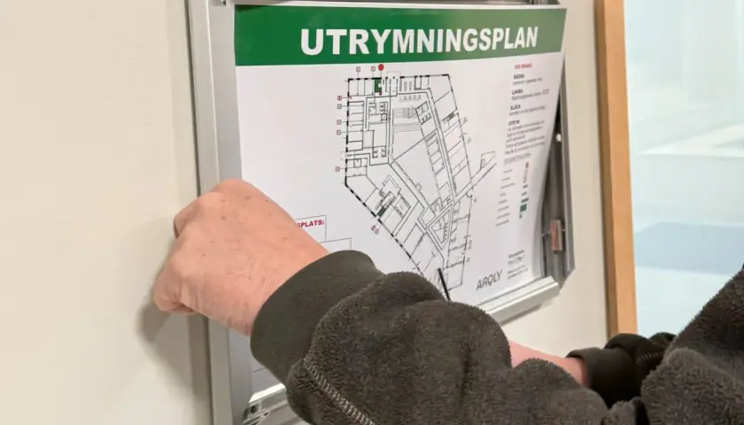 En hand justerar en utrymningsplan på väggen. Planen visar byggnadens planlösning och instruktioner för utrymning, vilket är viktigt enligt utrymningslarm regler.