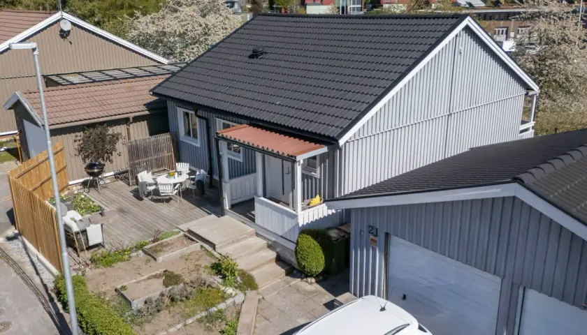 Ett charmigt hus med nummer bbr 21. Trädgårdsmöbler på altanen, en grill och en vit bil framför garaget.