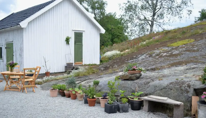 En vit stuga med gröna detaljer och ett bord med stolar på en grusad uteplats. Blommor och växter i krukor pryder den steniga sluttningen.
