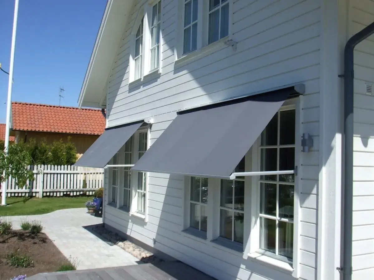 Grå markiser monterade på ett vitt trähus ger skugga. En vacker trädgård med gångväg syns i bakgrunden.