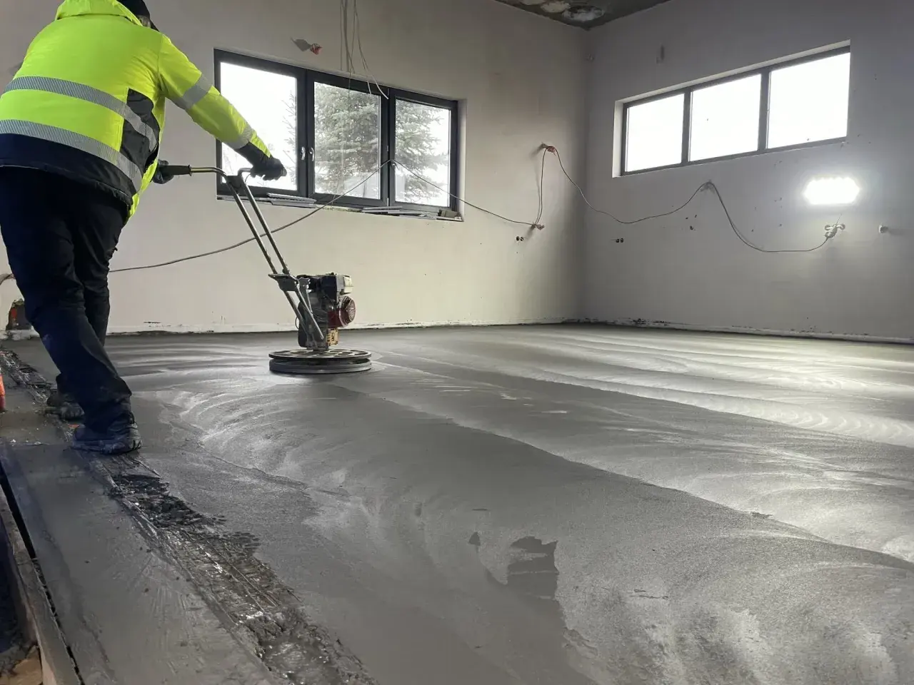 Arbetare polerar nylagd betong med en maskin. Golvet är fortfarande fuktigt och reflekterar ljuset. En del av ballast betong syns längs kanten.