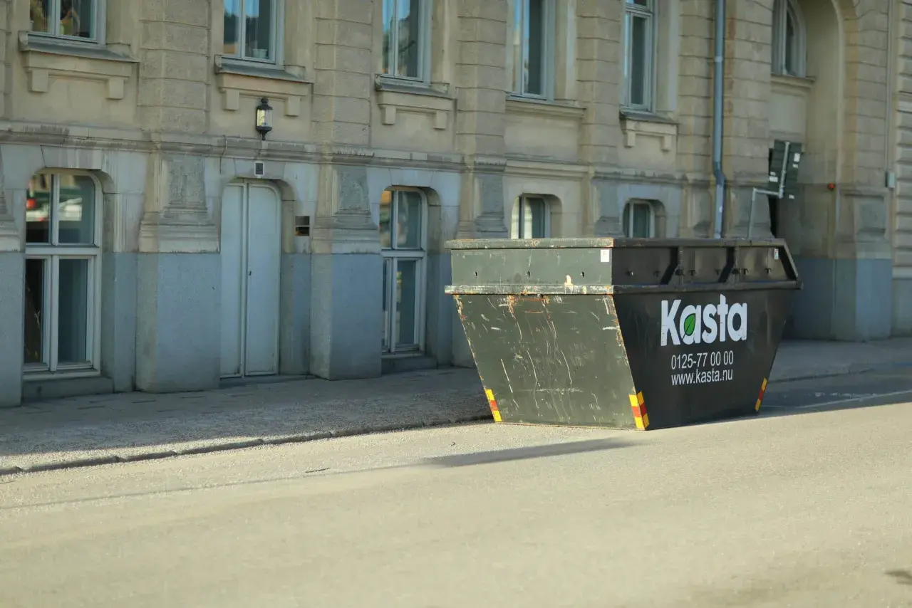Stor container för avfall står på gatan framför en byggnad. Den är märkt med "Kasta" och kontaktuppgifter.