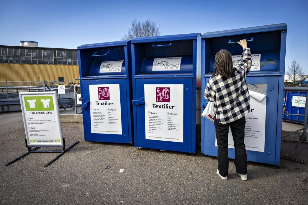 Person lämnar kläder i blå container för textilier återvinning. Skylt visar vad som får lämnas.