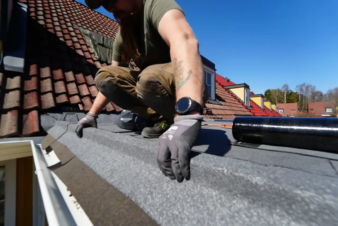 Man spikar takpapp på ett tak med tegelpannor. Solen skiner och himlen är blå.