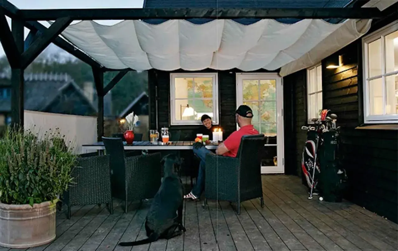En mysig uteplats med segelduk som tak. Ett par sitter vid ett bord med en hund, redo för en trevlig kväll.