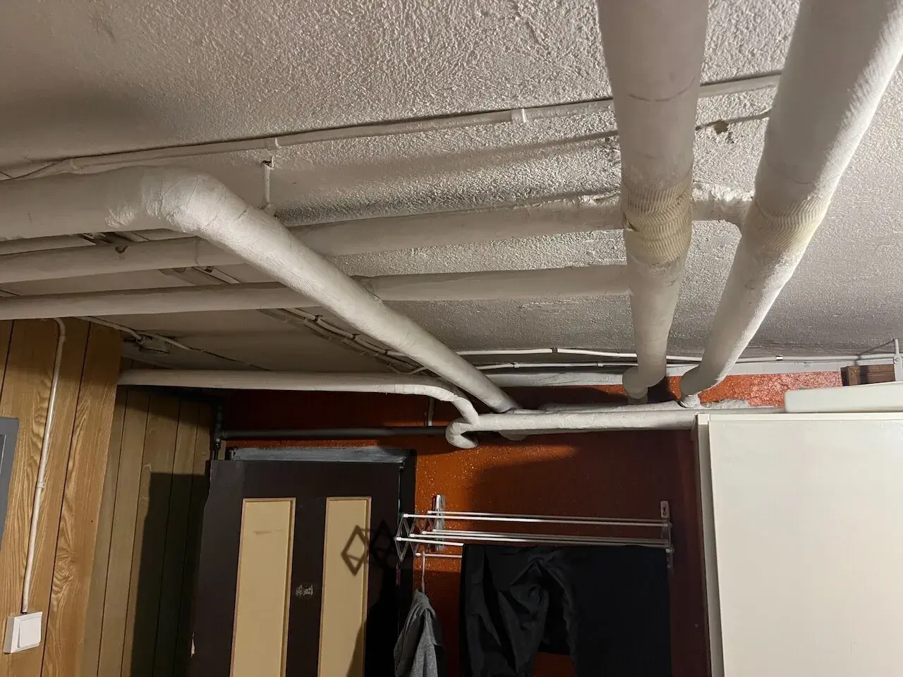 Isolerade rör löper längs det vitmålade svall taket i ett utrymme med träpanel och en dörr.