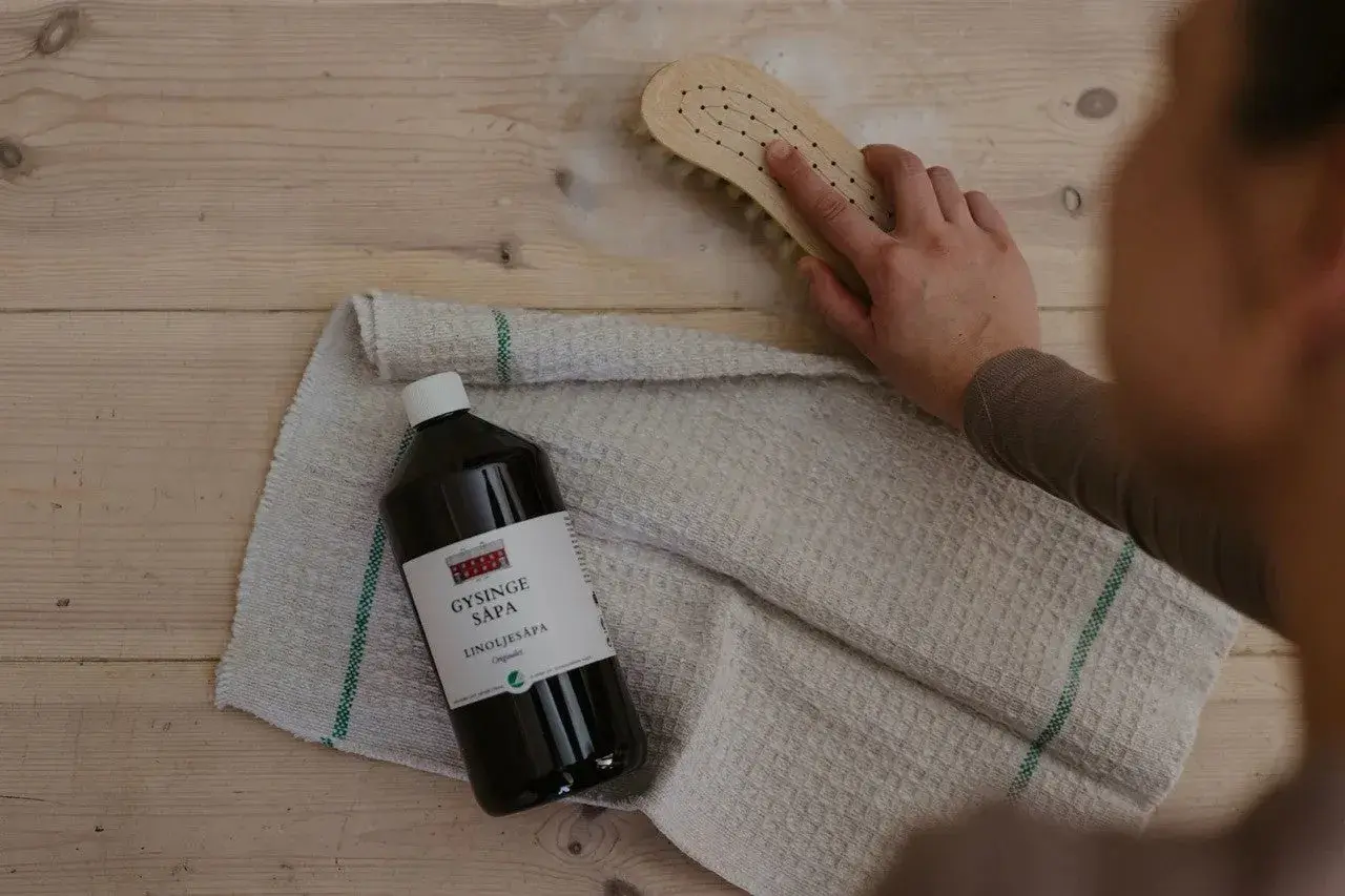 En hand skrubbar trägolv med en borste och linoljesåpa. Flaskan med linoljesåpa är märkbar, men var försiktig, den är brandfarlig.