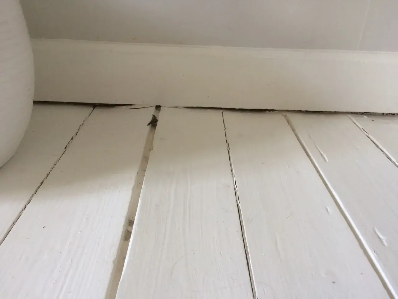 Vita träplankor med sprickor och ojämnheter, redo att slipa målat golv för en jämnare yta.
