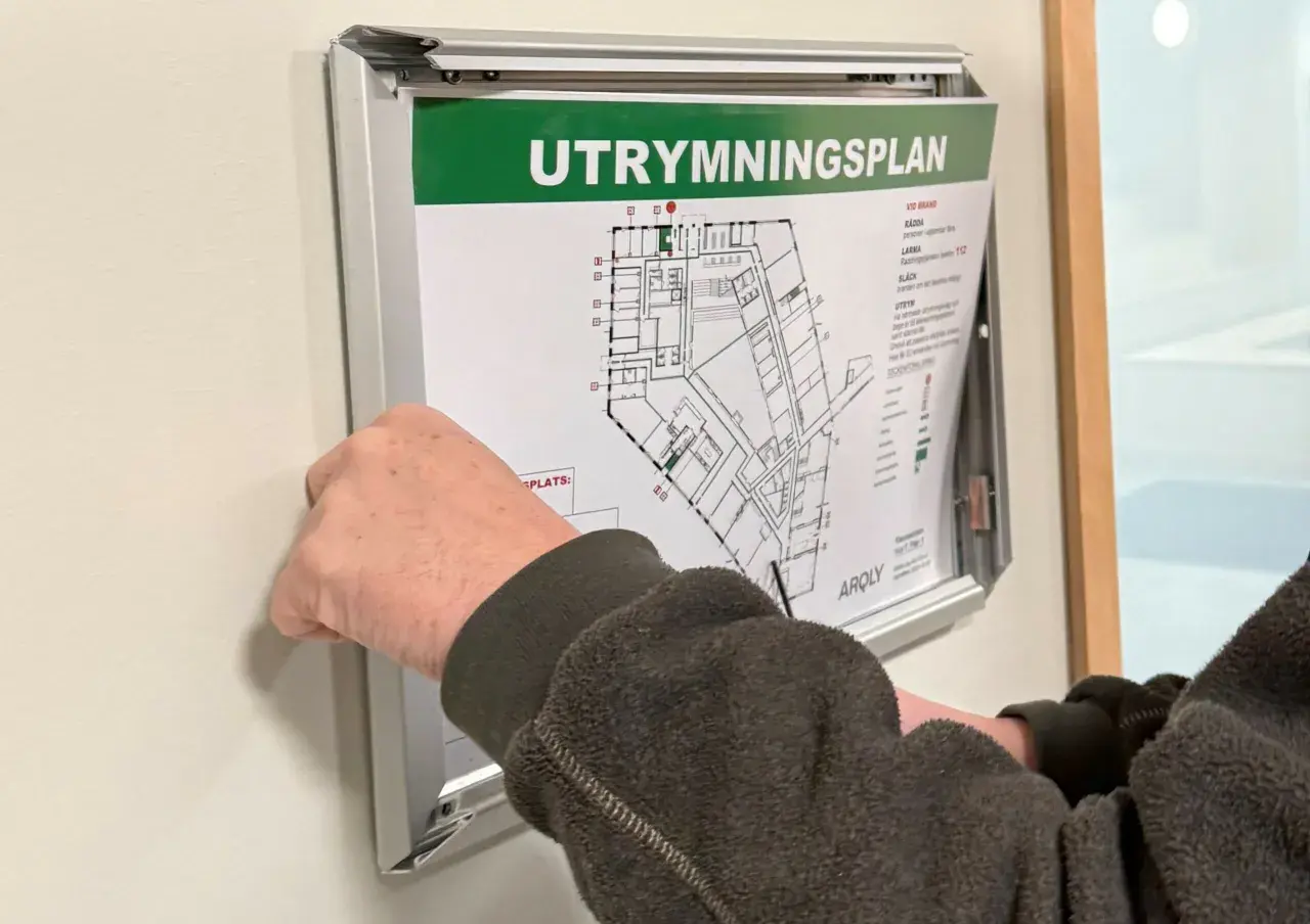 En hand justerar en utrymningsplan på väggen. Planen visar byggnadens planlösning och instruktioner för utrymning, vilket är viktigt enligt utrymningslarm regler.