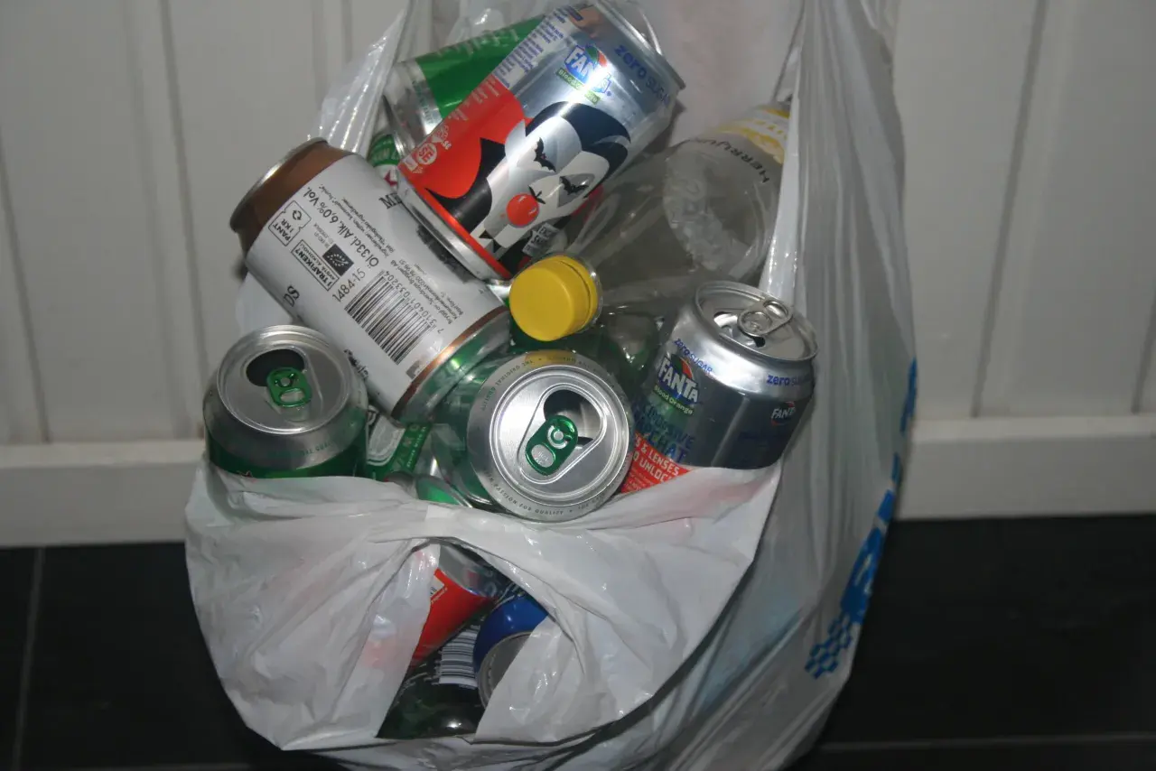 En plastpåse fylld med tomma burkar och flaskor, redo för pant. Här finns bland annat Fanta, ölburkar och en flaska med gul kork.