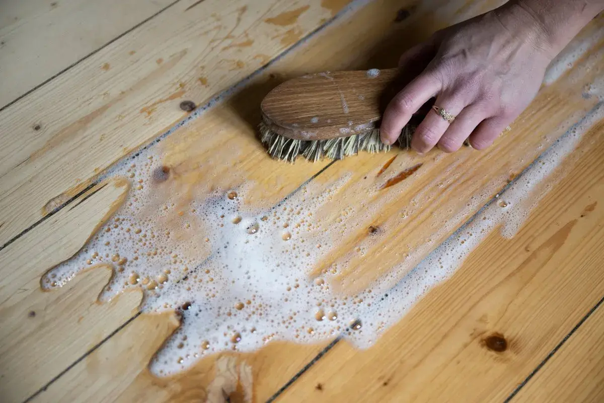 En hand skurar ett trägolv med en borste och såpvatten. Ett enkelt såpvatten recept kan ge ett skinande rent resultat.