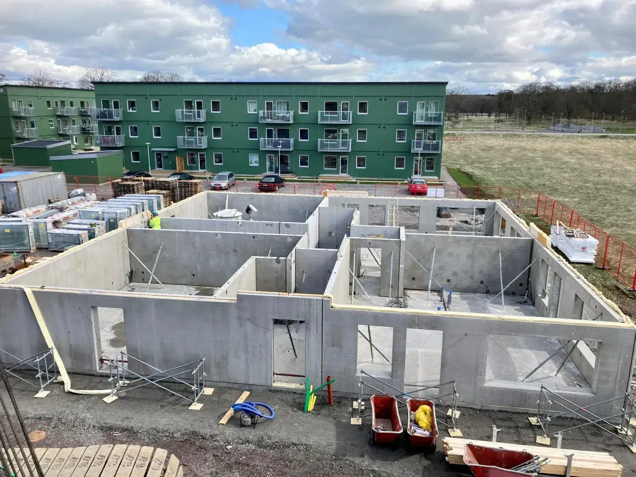 Byggarbetsplats med prefabricerade betongelement som formar grunden till ett hus. I bakgrunden syns gr&ouml;na flerfamiljshus.