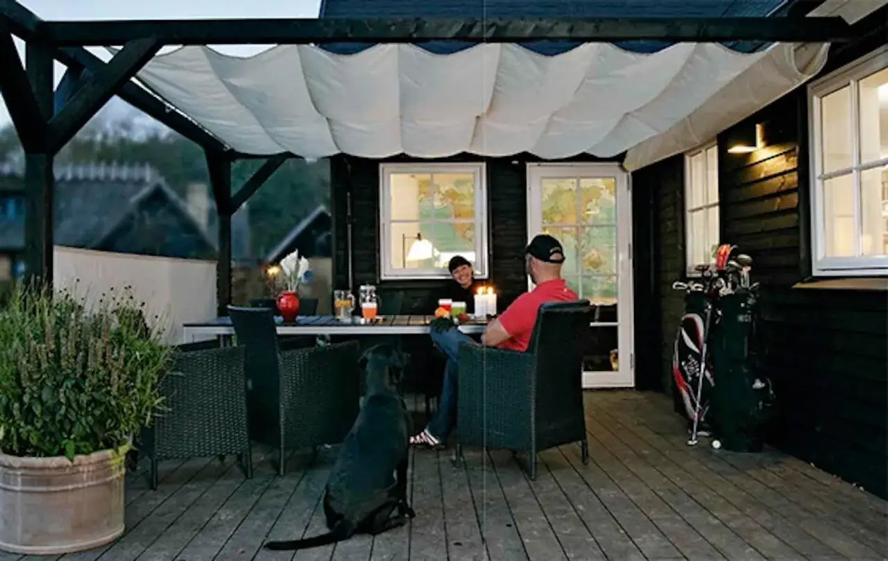En mysig uteplats med segelduk som tak. Ett par sitter vid ett bord med en hund, redo f&ouml;r en trevlig kv&auml;ll.