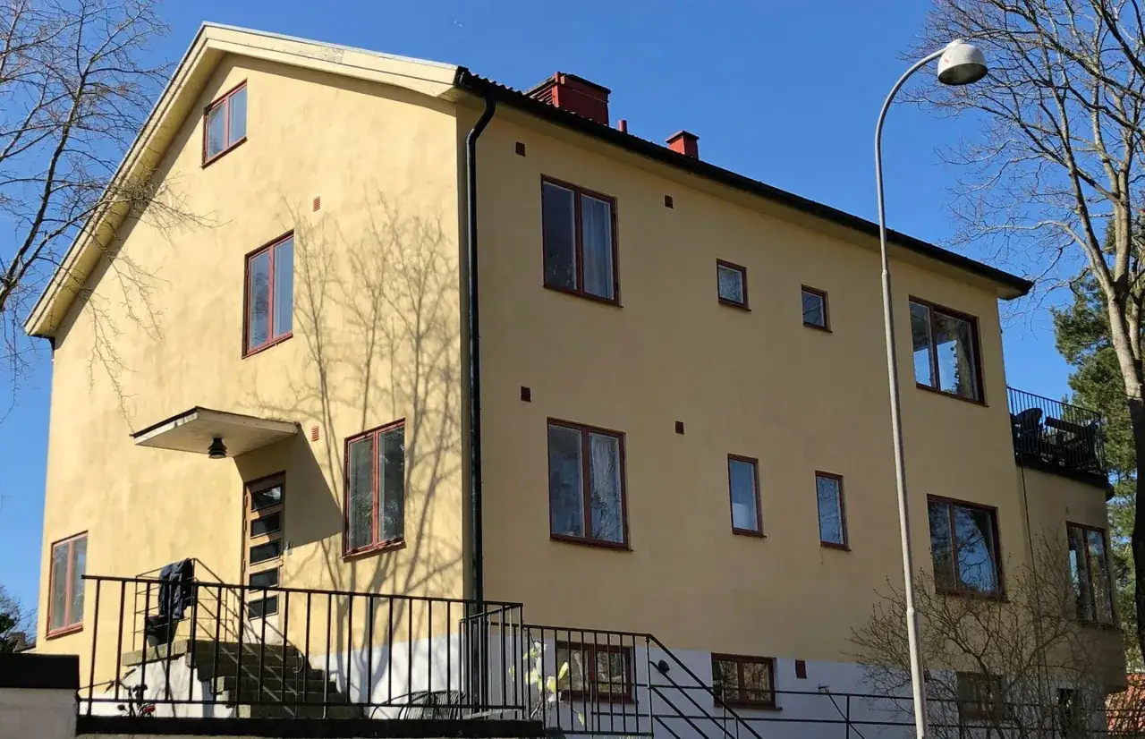 Gult funkishus fr&aring;n 40-talet med m&aring;nga f&ouml;nster och en balkong. Tr&auml;dens skuggor dansar p&aring; fasaden.