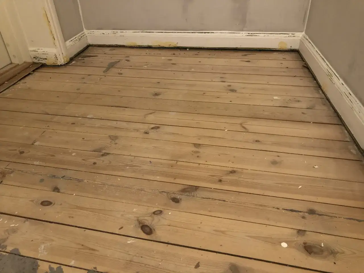 Obehandlat tr&auml;golv av gran eller furu med synliga kvistar och sp&aring;r av f&auml;rg. Golvet &auml;r i ett h&ouml;rn med vita golvlister.